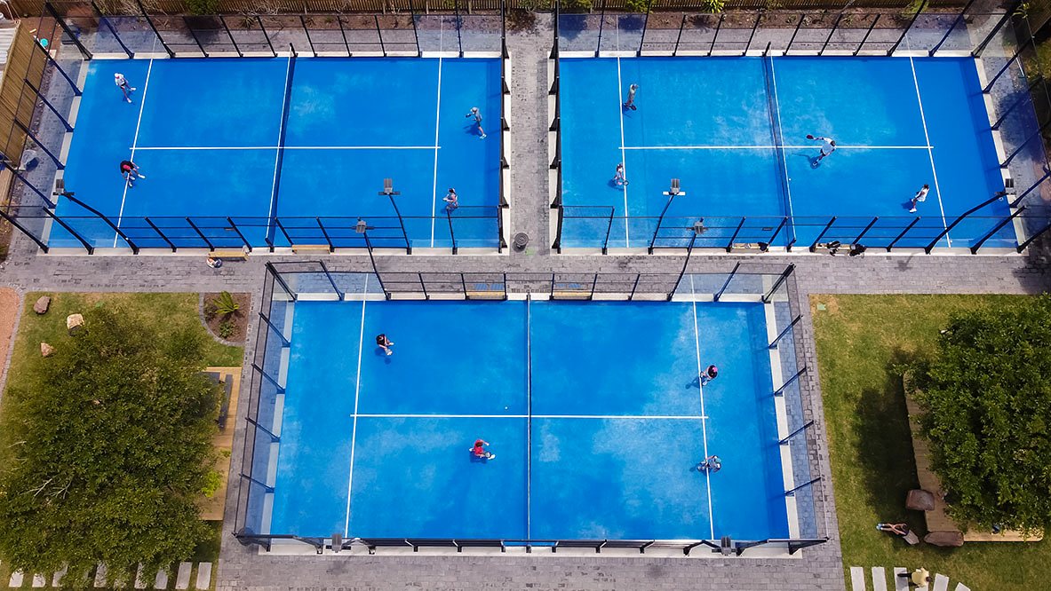 Three blue padel courts at the River Club seen from above, with games in progress