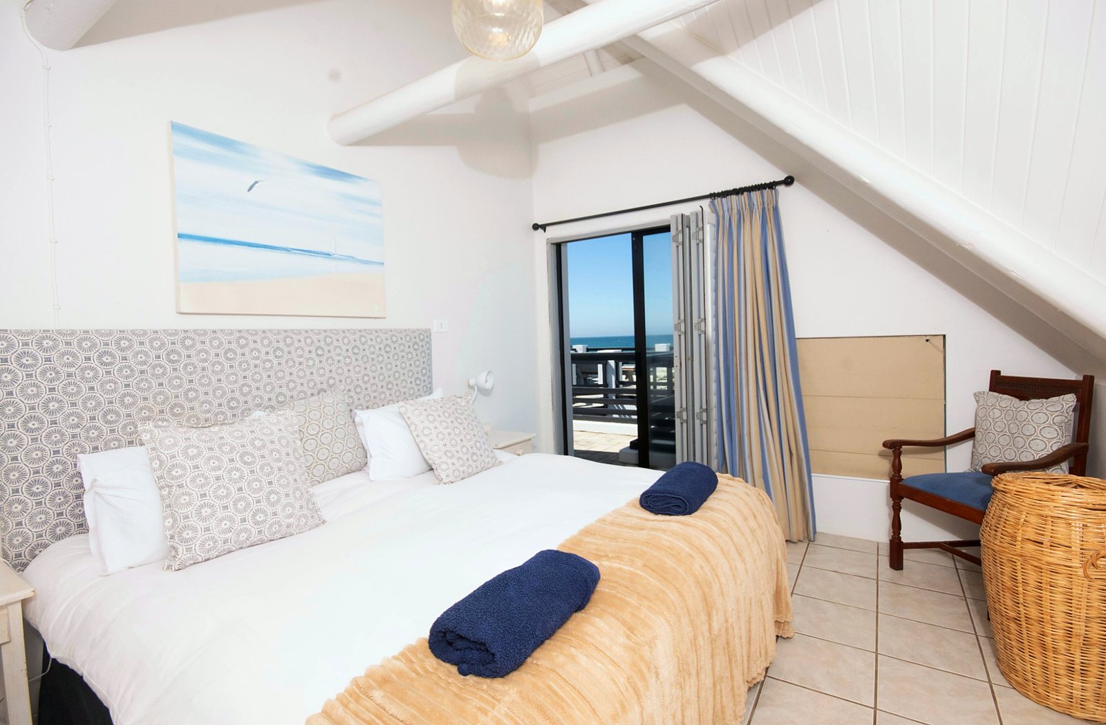 Bedroom with a king bed under a pitched white ceiling, the door open to a balcony and the sea